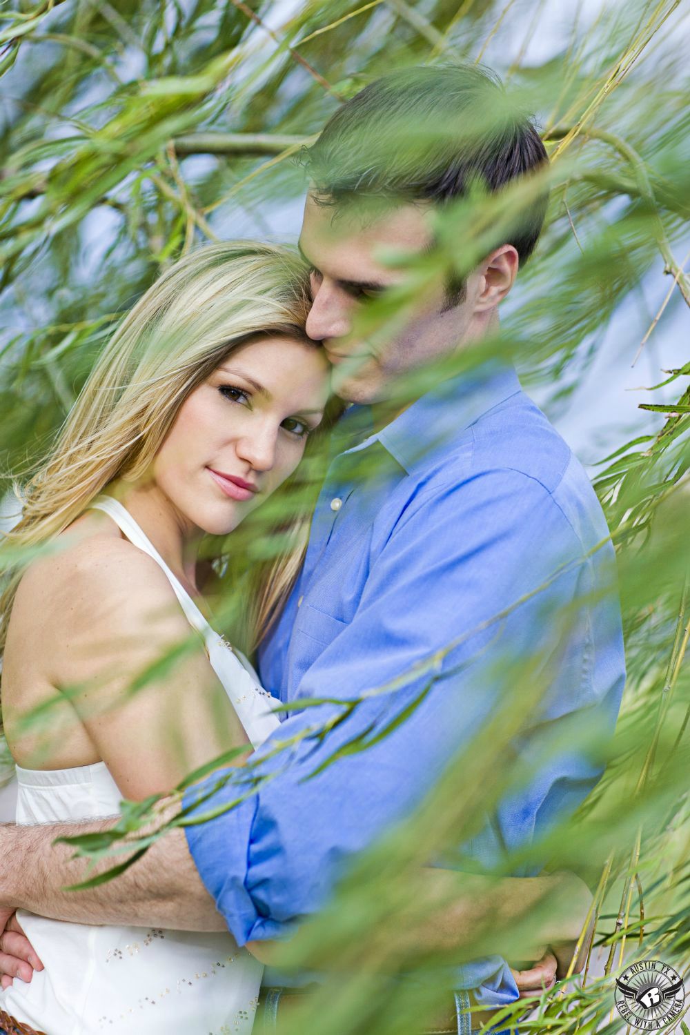 Content, beautiful blond girl wearing white halter top shirt embraces a dark haired guy wearing a blue button up dress shirt surrounded and obscured by willow tree branches with green leaves at Butler Park near the pond in this charming engagement photo in downtown Austin.